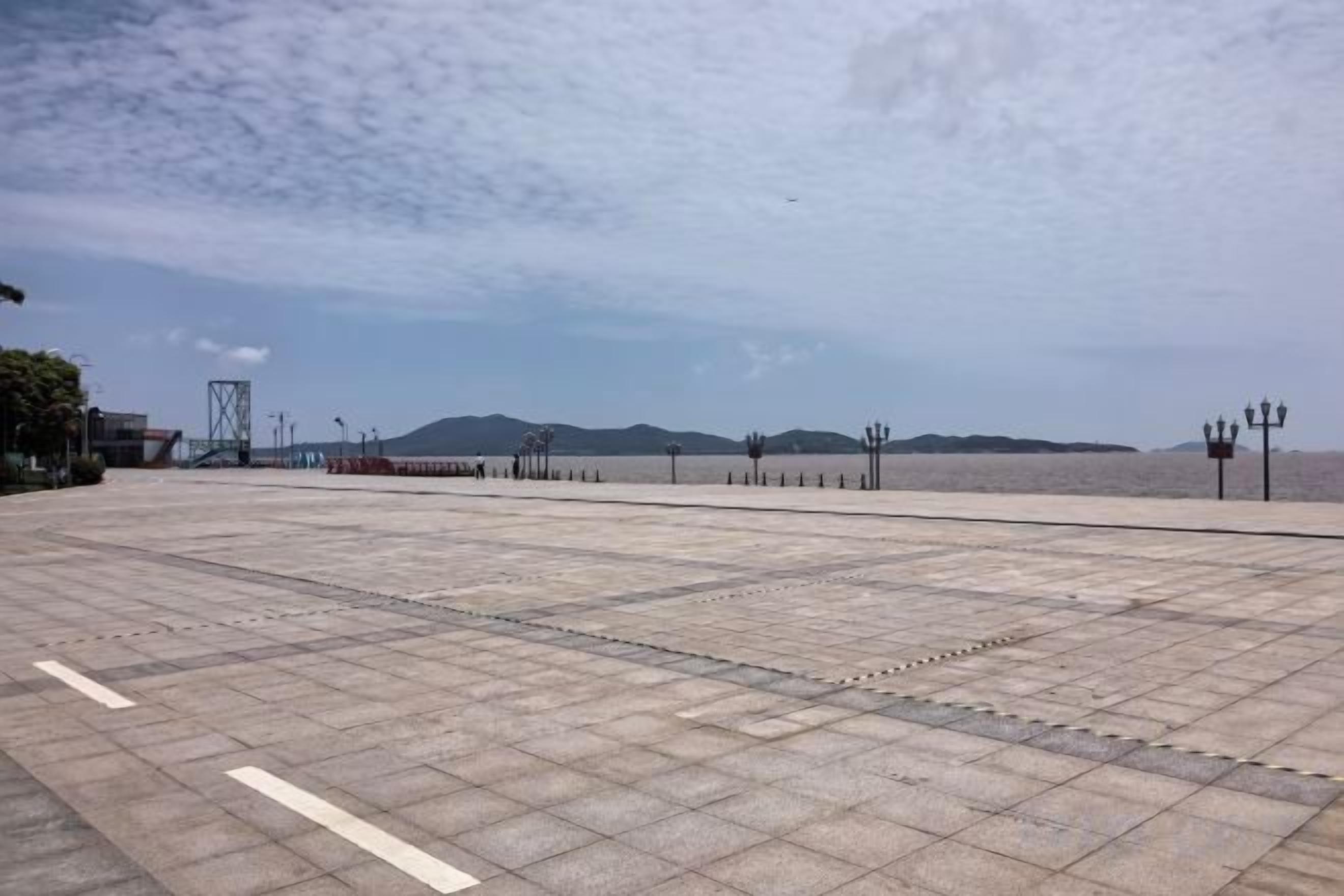 A paved waterfront promenade with a clear sky, distant mountains, and street lamps.