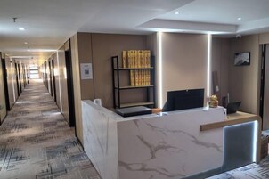 Zona de recepción de un hotel con un mostrador de mármol, una estantería y un largo pasillo.