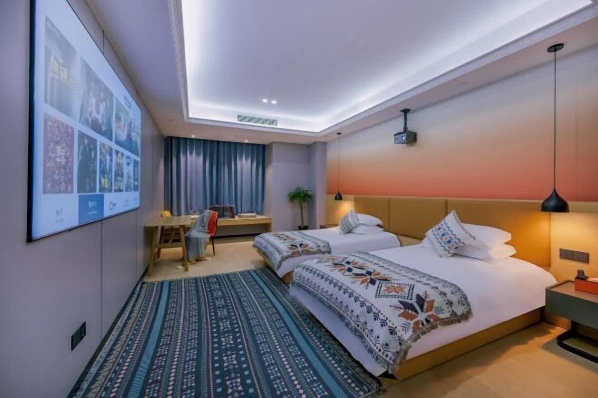 Habitación de hotel con cama, mesitas de noche, escritorio, silla, planta y un cartel grande en la pared.