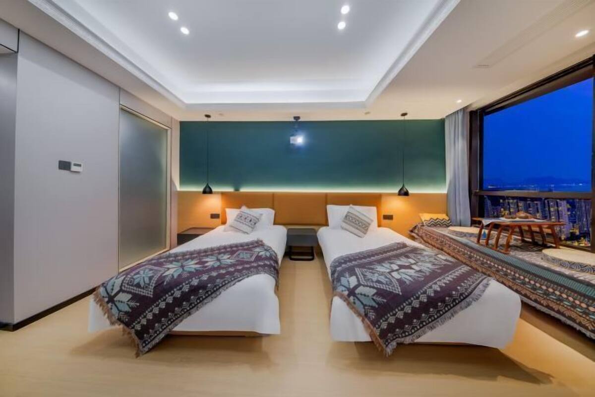 Una habitación de hotel moderna con dos camas, un ventanal con vistas a la ciudad y una zona de estar con mesa y sillas.