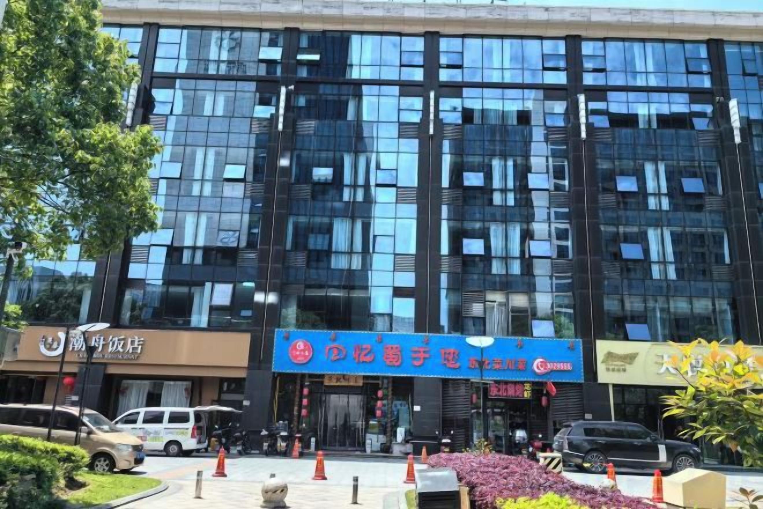 A modern building with a glass facade, a blue sign with white text, and a parking lot with cars and traffic cones.