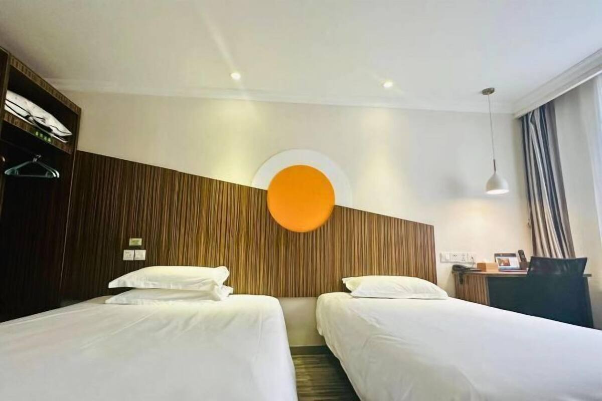 Een hotelkamer met twee bedden, een accentmuur van hout, een ronde oranje lamp, een bureau met stoel en een raam met gordijnen.