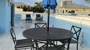 Eine Dachterrasse mit rundem Tisch und Stühlen, einem blauen Sonnenschirm und einem Pool im Hintergrund.