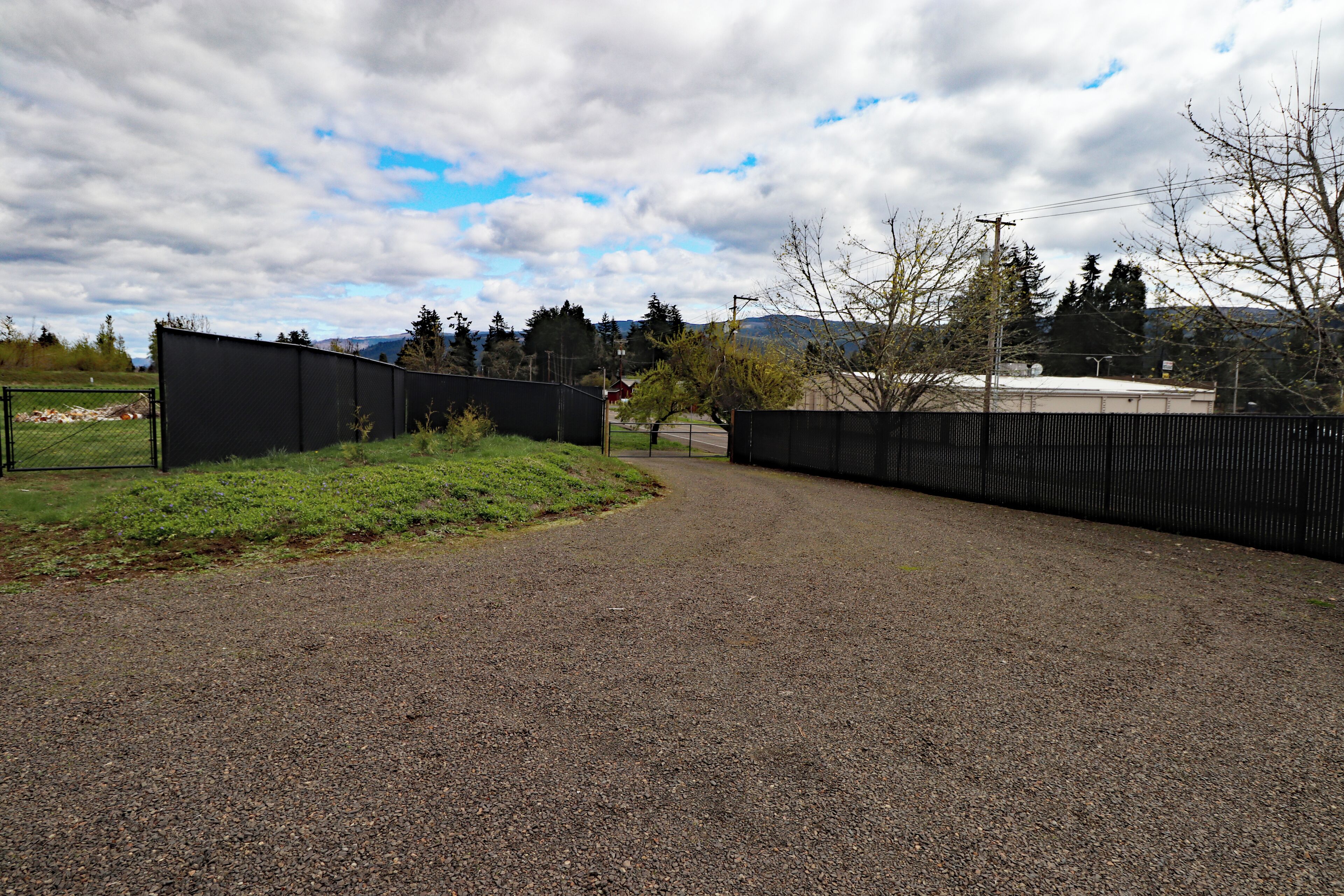 A gravel road leading to a gated area with a fence, surrounded by greenery and trees.