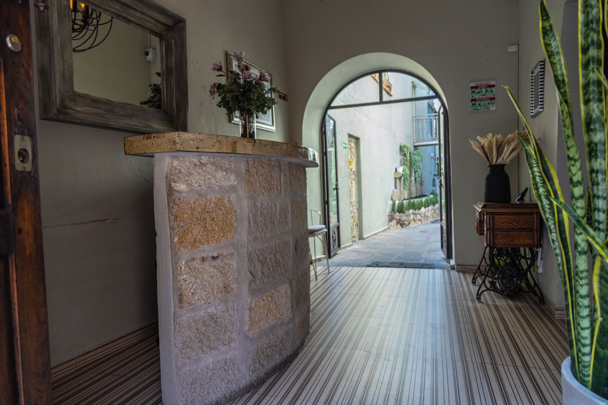 A hallway with a stone reception desk, a mirror, a potted plant, and an arched doorway leading to an outdoor area.