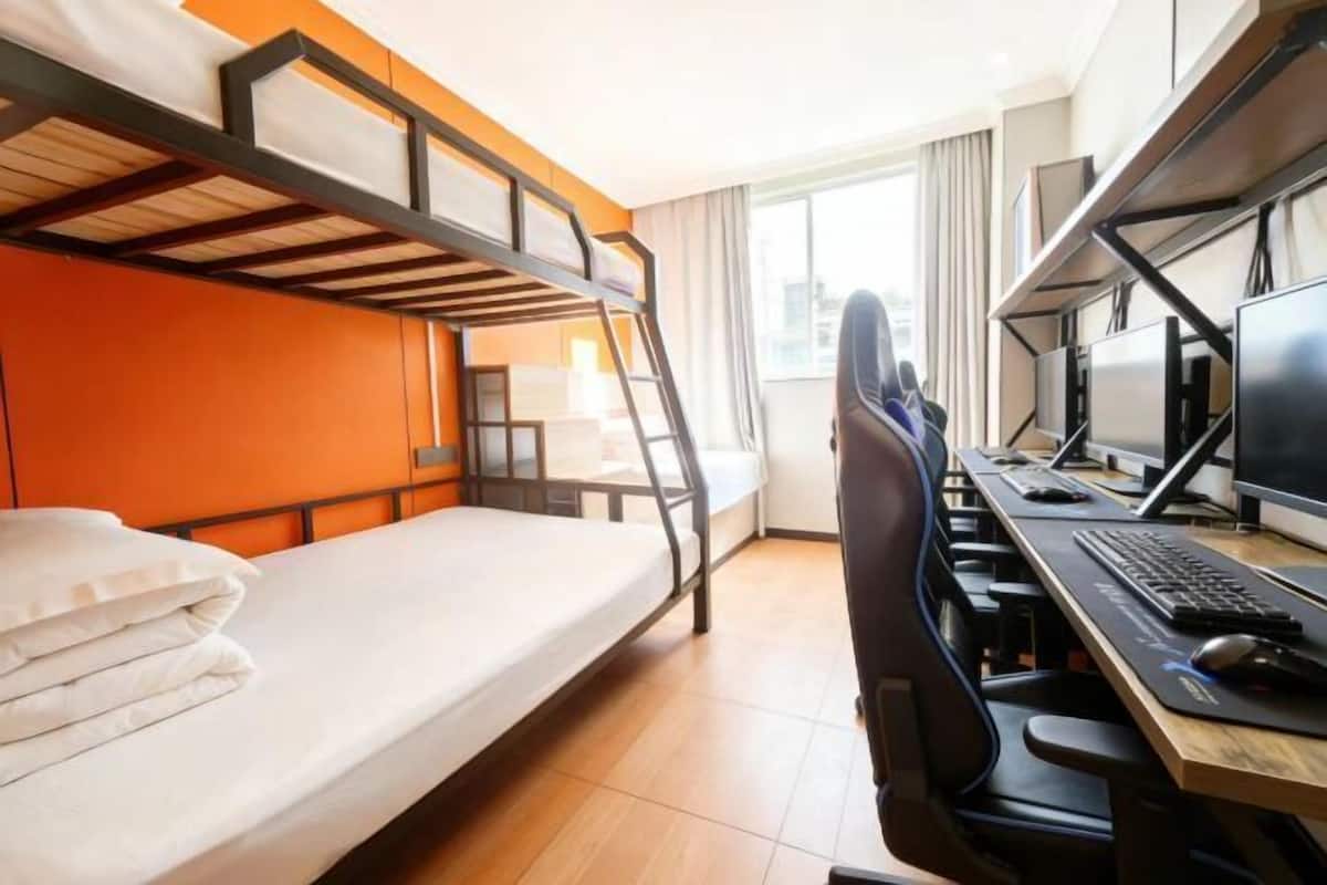 Uma cama beliche com escrivaninha e computador, janela com cortinas e vista para a cidade.