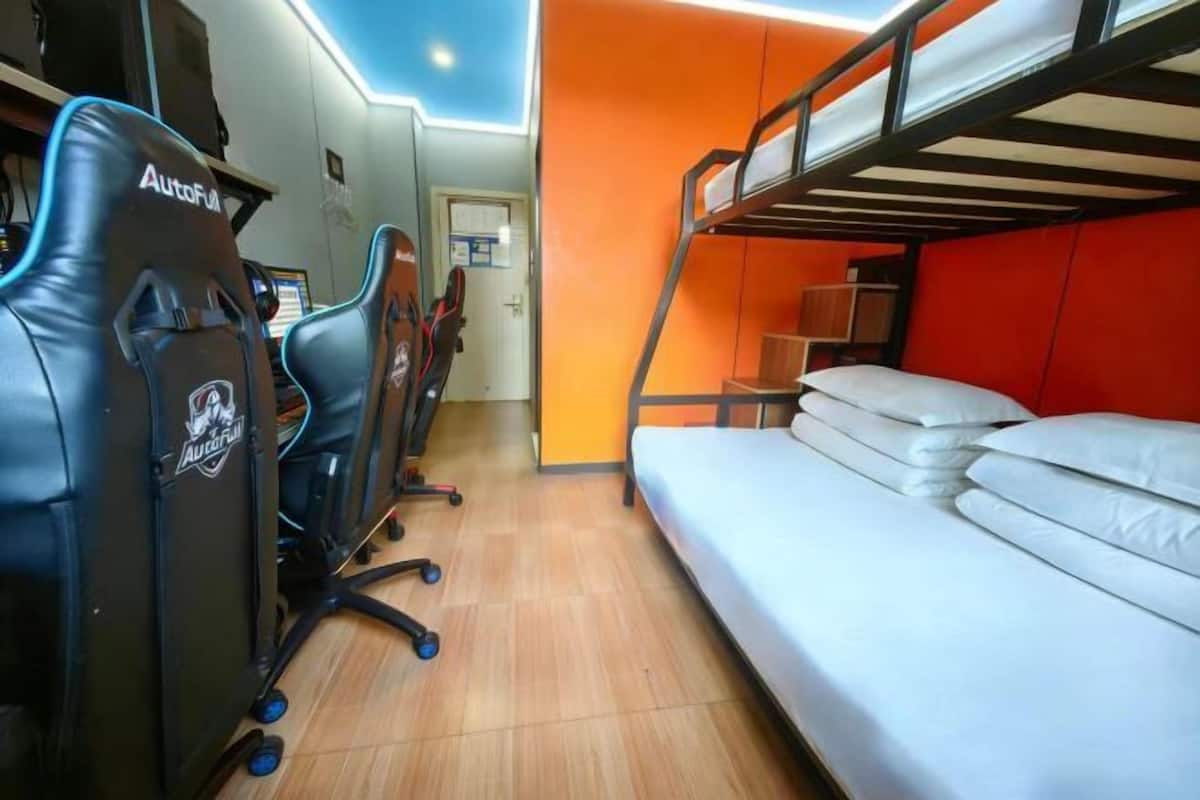 Uma cama beliche com colchão branco, uma escrivaninha com duas cadeiras gamer pretas e um piso de madeira.