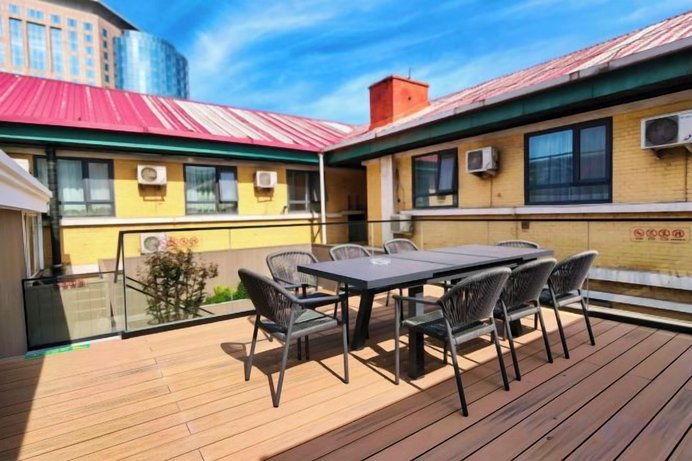 Une terrasse extérieure avec une table et des chaises, une balustrade en verre et un bâtiment doté d’un toit rouge et de climatiseurs.