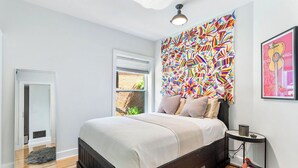 Une chambre à coucher moderne avec un grand lit, une œuvre d’art abstraite aux couleurs vives accrochée au mur, une petite table d’appoint et une chaise.