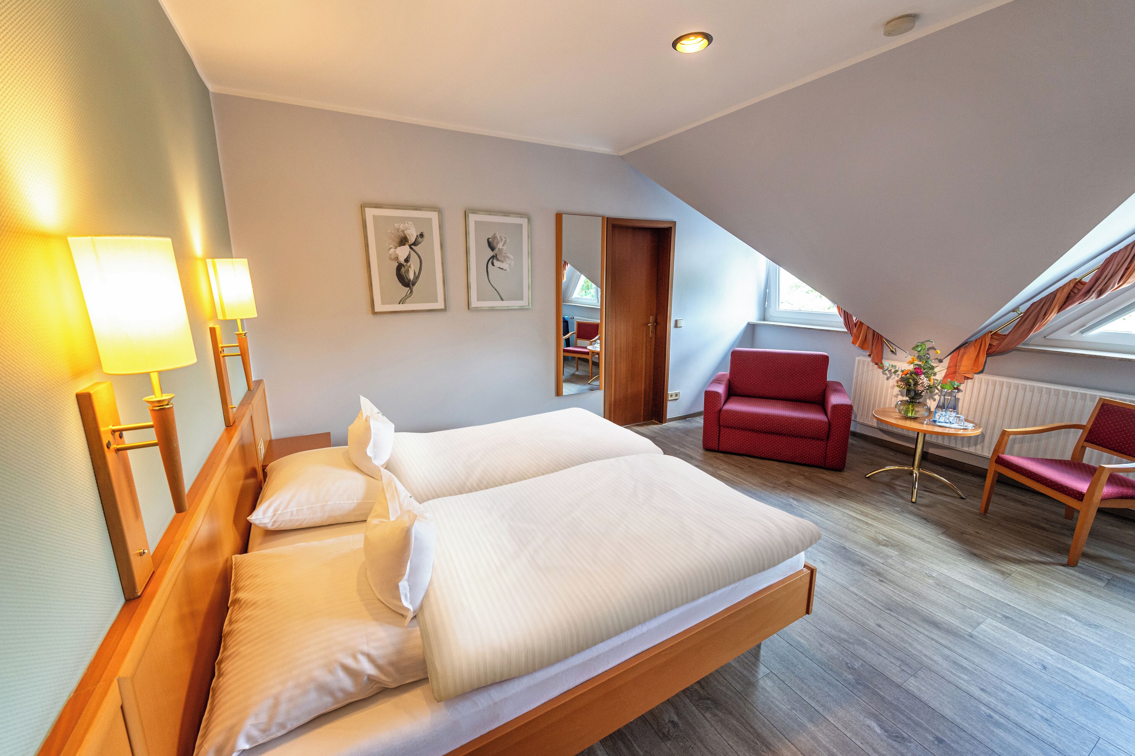 Ett hotellrum med en säng, två sänglampor, en röd fåtölj, ett träbord och en spegel.