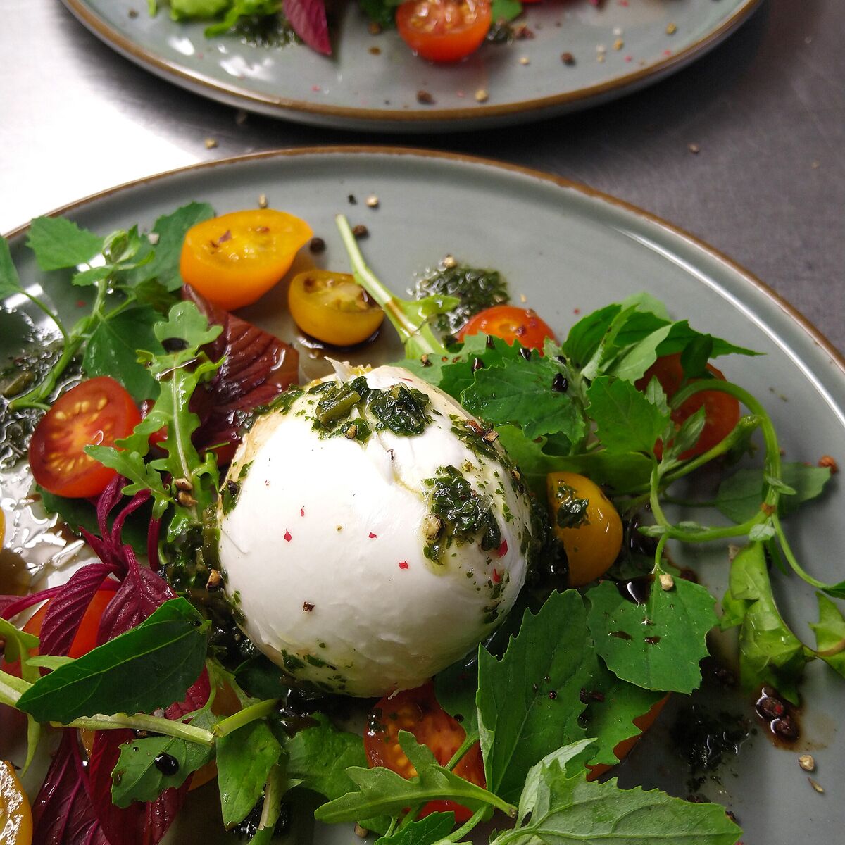 Ein Teller mit Essen, darunter eine weiße, kugelförmige Zutat, möglicherweise Mozzarella, umgeben von Grünzeug und Kirschtomaten.