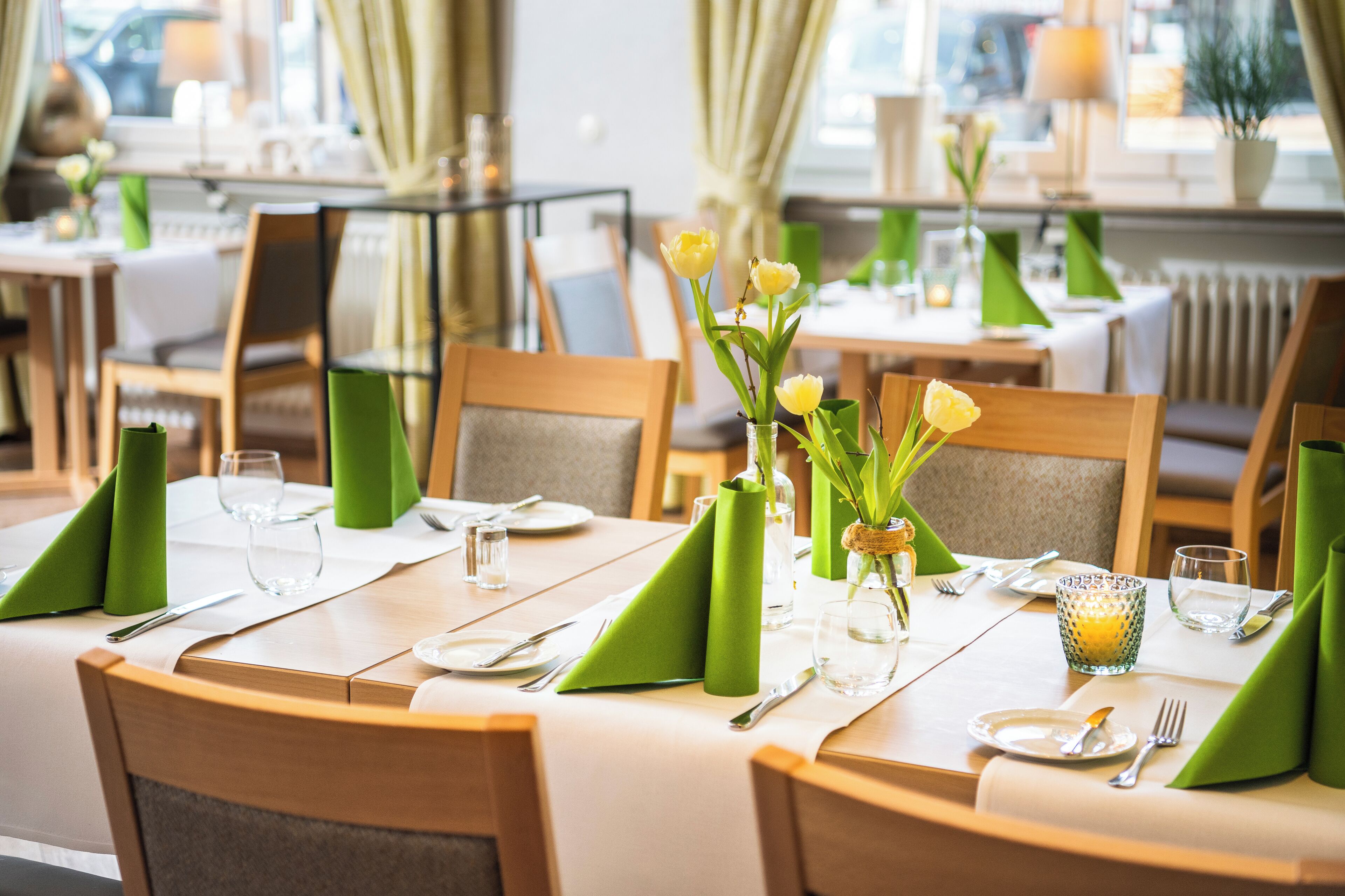 Un área de comedor con mesas dispuestas para dos, cada una con servilletas verdes, cubiertos y un jarrón con flores amarillas.