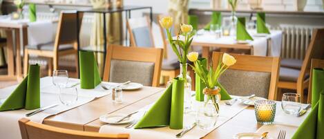 Un área de comedor con mesas dispuestas para dos, cada una con servilletas verdes, cubiertos y un jarrón con flores amarillas.