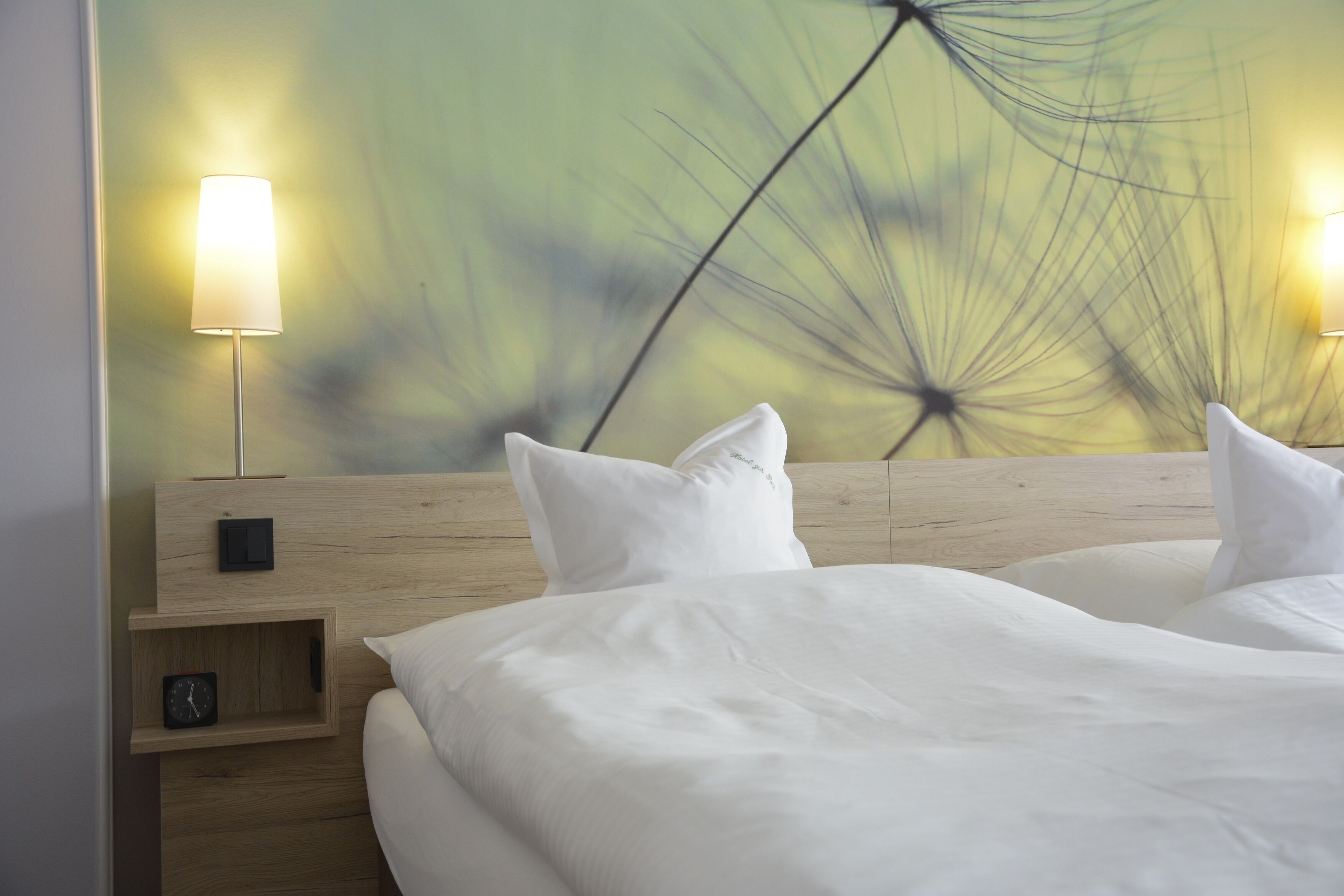 Ein Hotelzimmer mit einem hölzernen Kopfteil, zwei Nachttischen mit Lampen, einer Uhr und einer Wand mit einem Löwenzahnmuster.