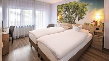 A hotel room with two beds, a desk with a chair, a large window with sheer curtains, and a wall mural of a tree.