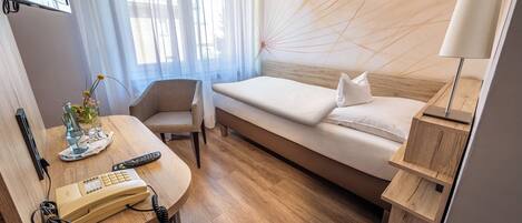 Ein Hotelzimmer mit einem Bett, einem Schreibtisch mit Telefon, einem Sessel, einer Lampe und einem am Wand montierten Fernseher.