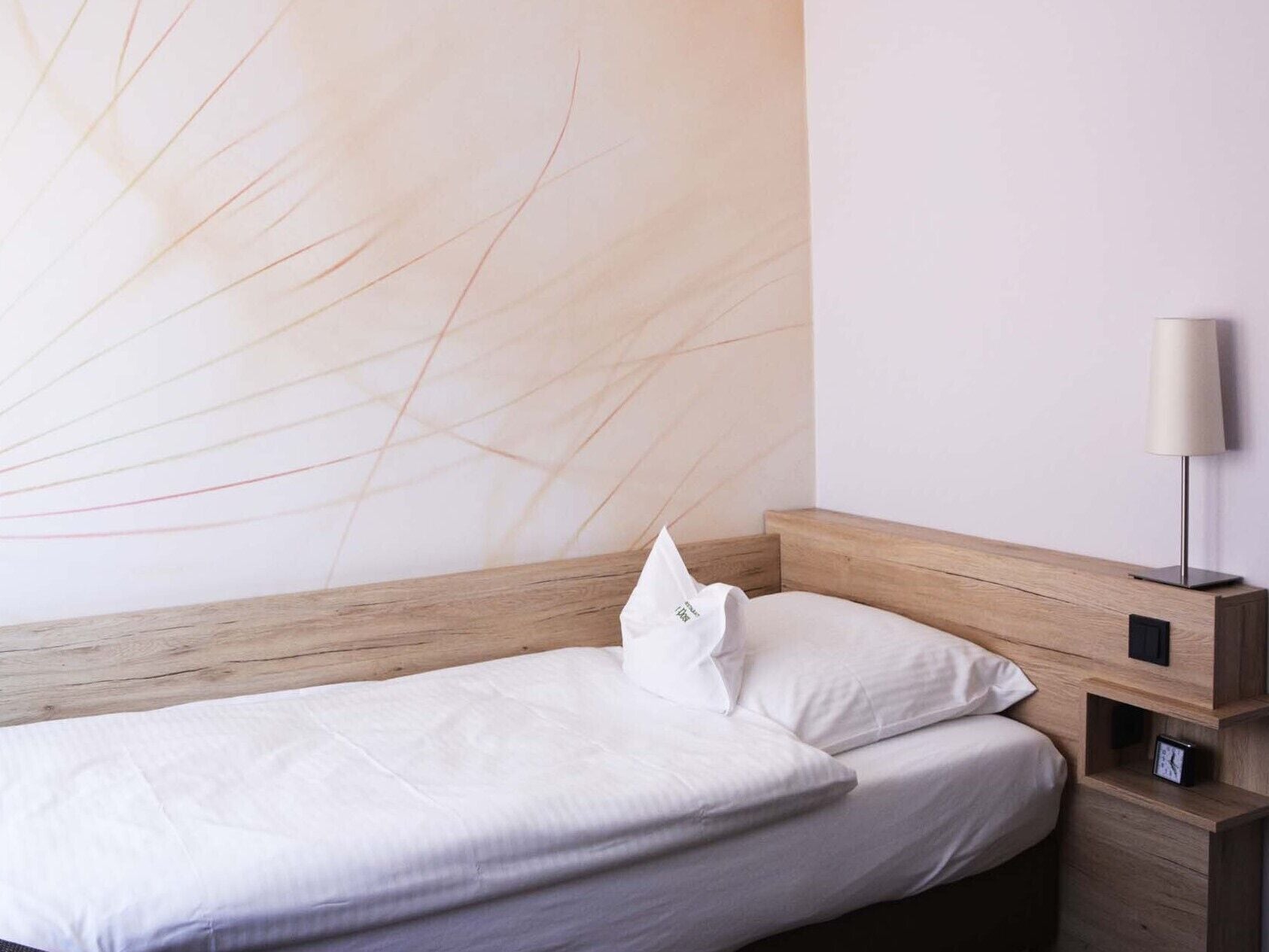 A single bed with a wooden headboard, white bedding, and a nightstand with a lamp and a clock.