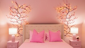 Ett sovrum med ett rosa tema, med en stoppad sänggavel, rosa sängkläder och sängbord med lampor och dekorativ belysning.