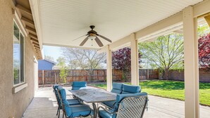 Un patio techado con una mesa y sillas, un ventilador de techo y vistas a un jardín trasero con cercado de madera y árboles.