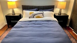 A neatly made bed with a grey headboard, two bedside tables with lamps, and a window with blinds.