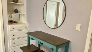 A room with a white dresser, a teal desk with two stools, and a mirror on the wall.