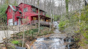Una cabaña roja con un balcón de madera y una cascada al fondo.