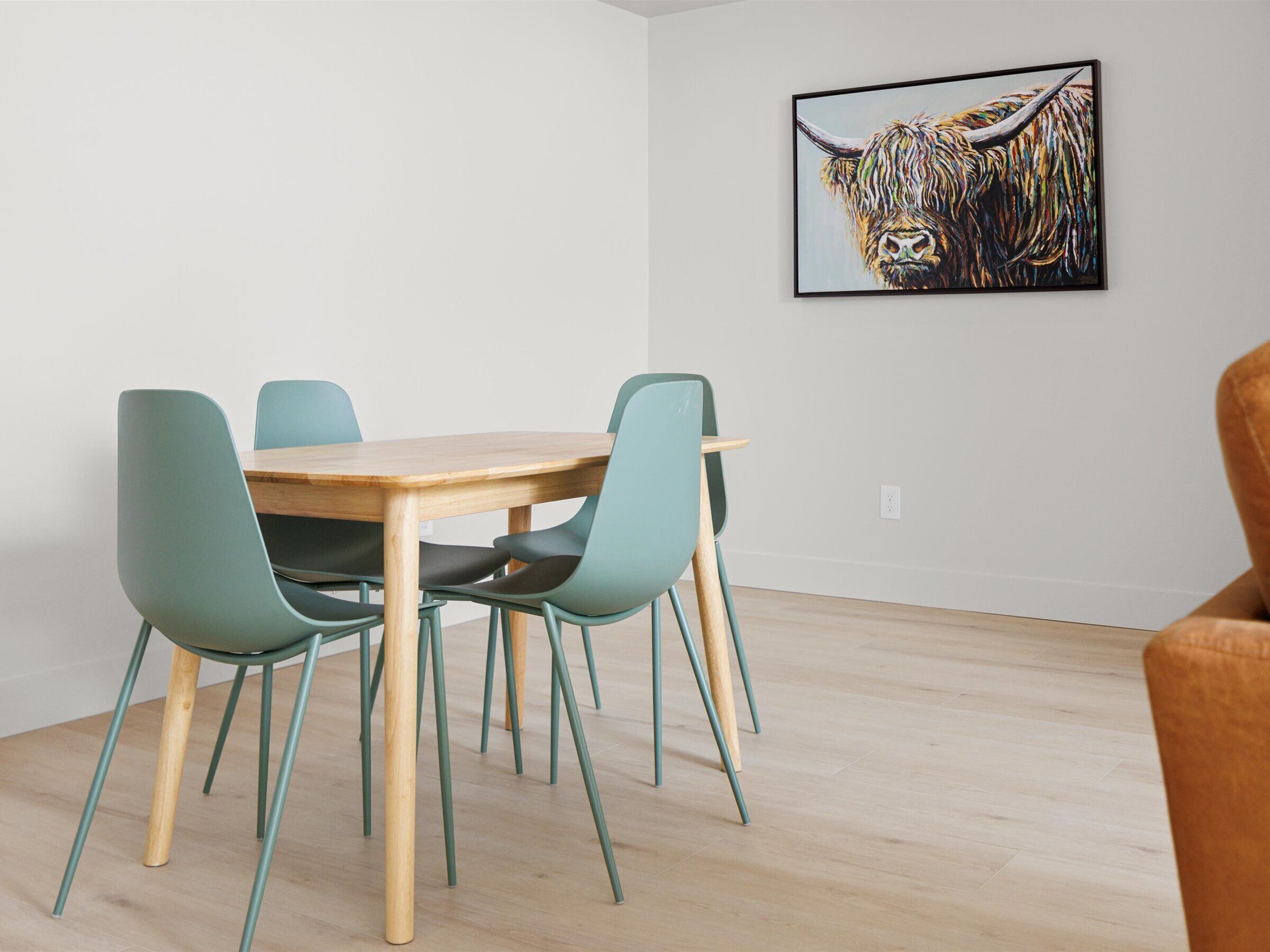 A dining area with a wooden table and four teal chairs. A framed artwork of a bull is on the wall.