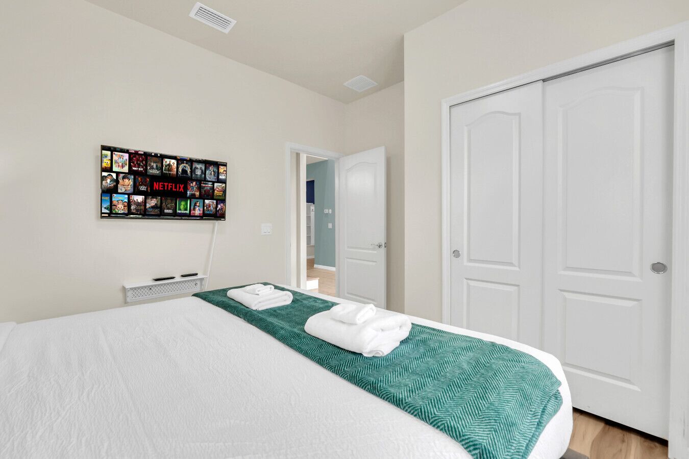 A hotel room with a neatly made bed, a wall-mounted TV displaying various shows, and a set of white doors.