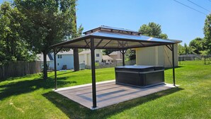 A covered outdoor hot tub area with a metal roof and a concrete base, surrounded by a grassy yard and a wooden fence. - Hawthorne Hideaway (West Des Moines)