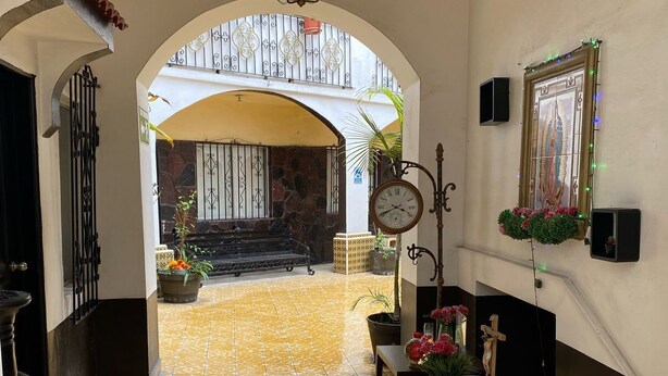 A hallway with yellow patterned tiles, a bench with flowers, a clock, and a framed religious image. - HOTEL YOLANDA (Tijuana)