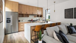 A modern kitchen with wooden cabinets, a stainless steel refrigerator, and a central island with bar stools. - The West Twin Oasis - Hot Tub near Downtown Austin (Austin)