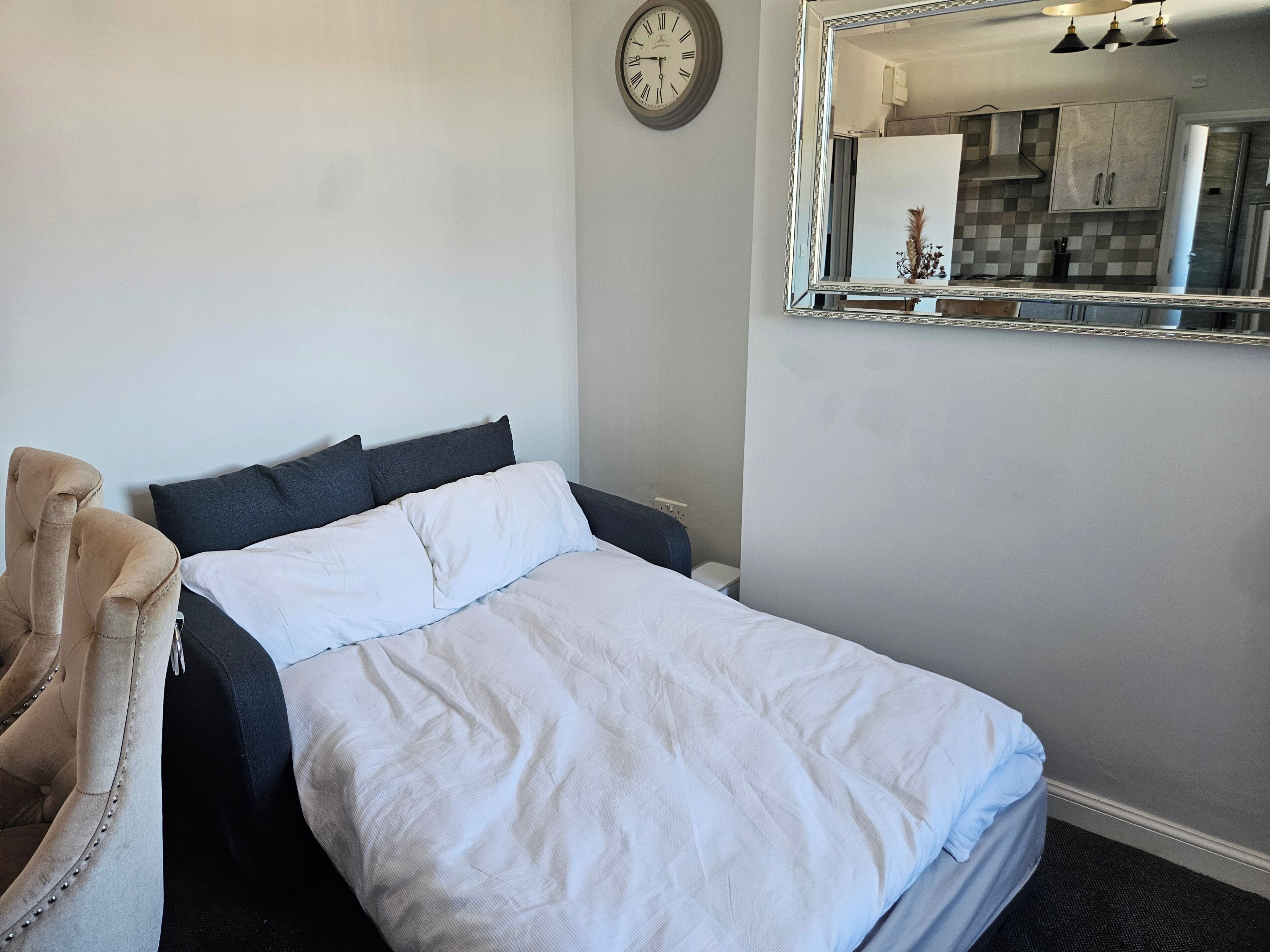 A neatly made bed with white and dark grey bedding, a round wall clock, and a kitchen visible in the mirror reflection.