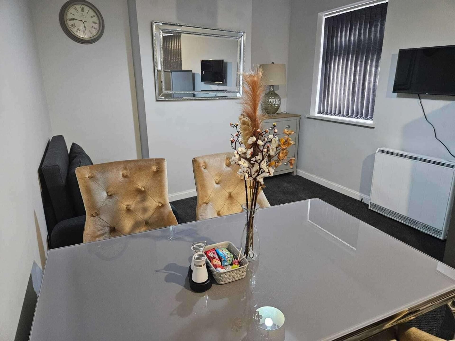 A conference room with a glass table, two upholstered chairs, a clock on the wall, a mirror, a radiator, and a TV mounted on the wall.
