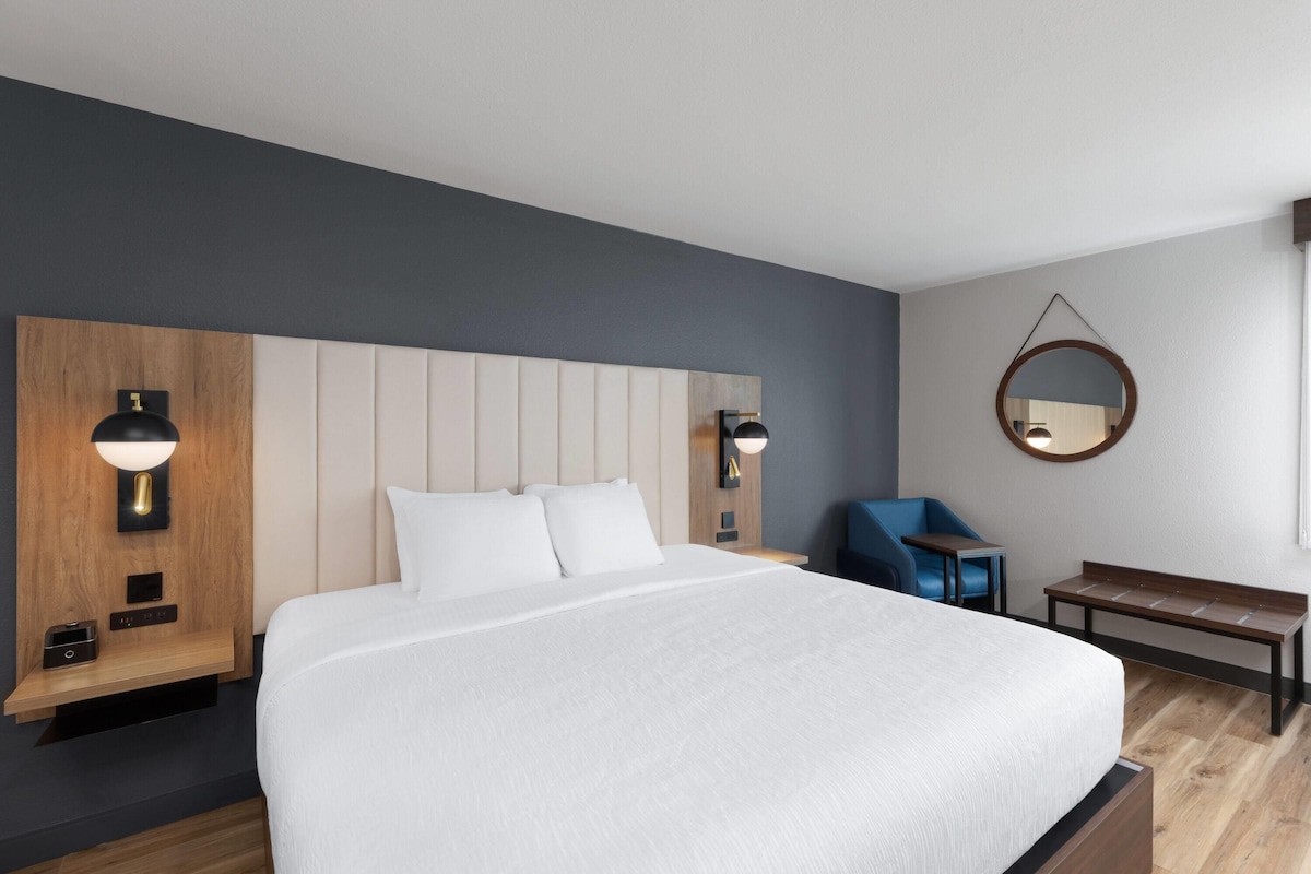 A modern hotel room with a large bed, bedside tables, a desk, a chair, and a round mirror on the wall.