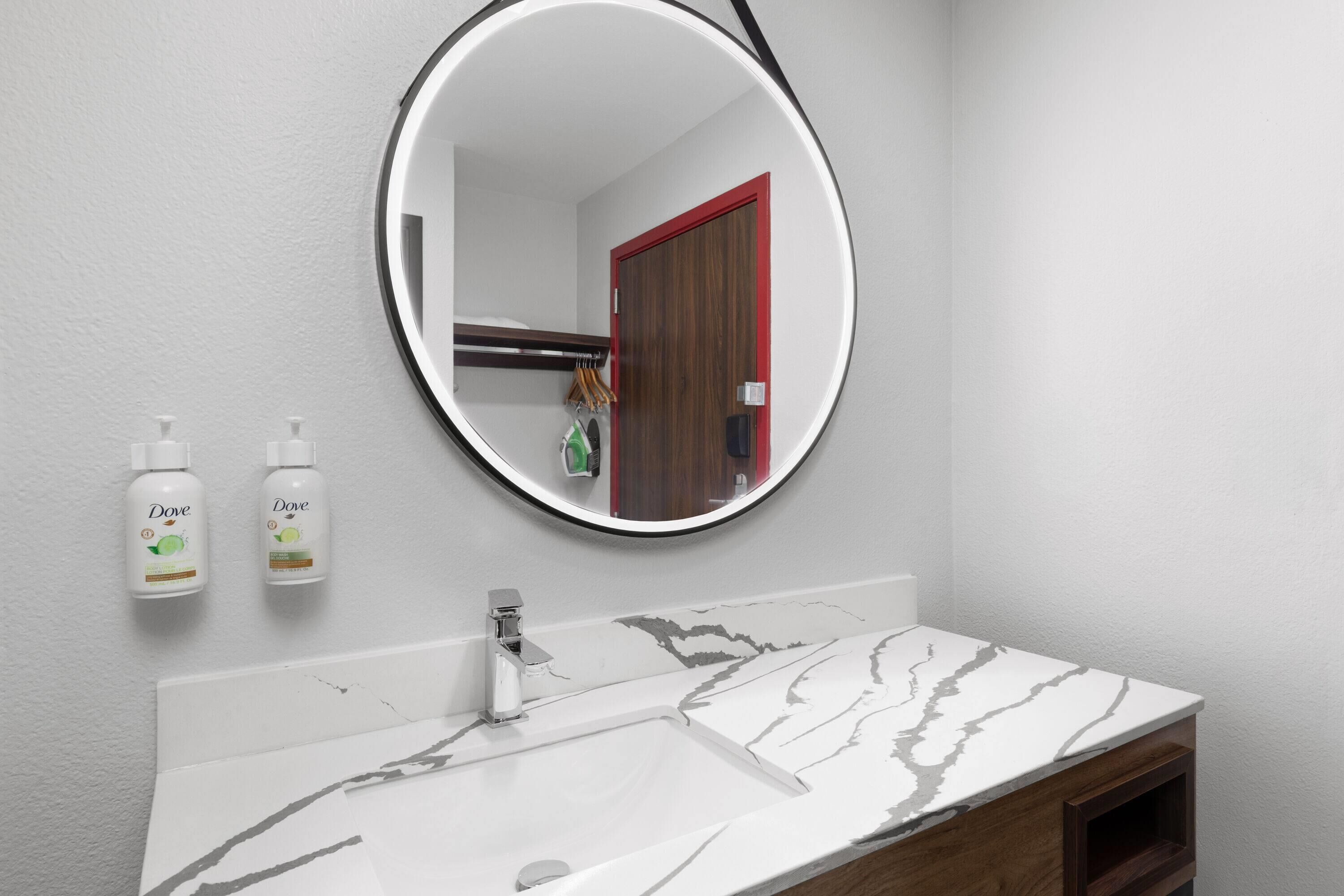 A bathroom with a white marble sink, a round mirror, and two Dove hand soap bottles.