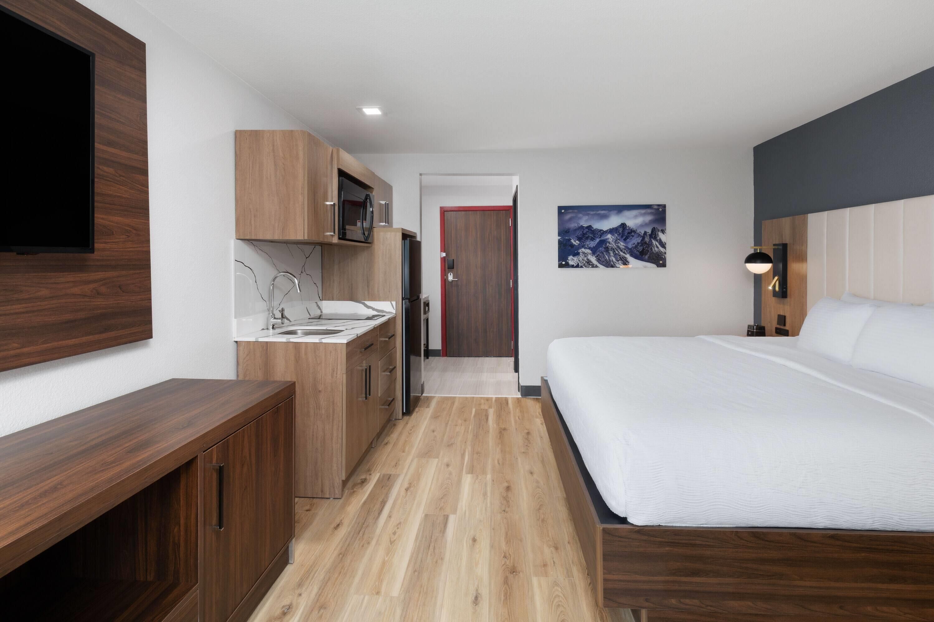 Une chambre d’hôtel moderne avec un plancher en bois, un lit avec une literie blanche, un téléviseur encastré, un coin cuisine avec des placards en bois et un tableau encadré au mur.