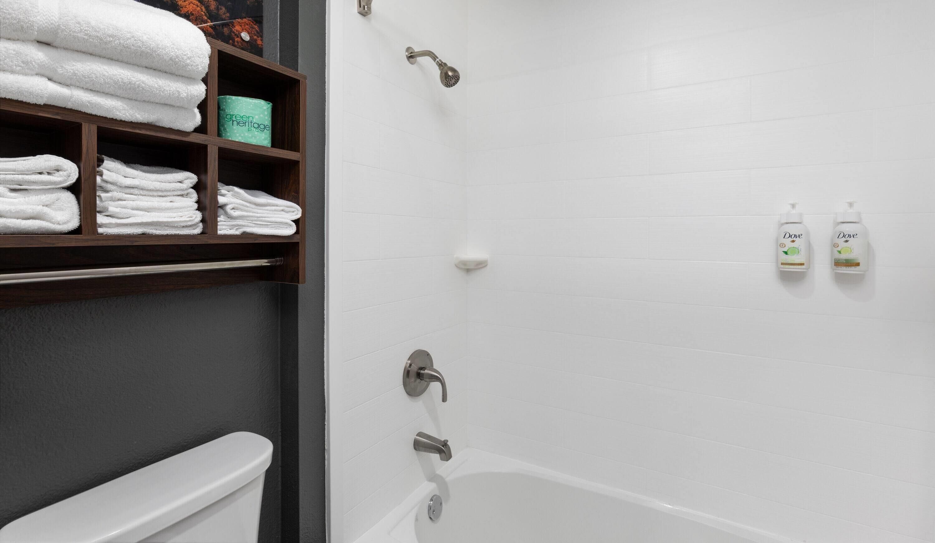 Un baño con una bañera blanca, un inodoro, una ducha y un estante con toallas dobladas y una botella verde.