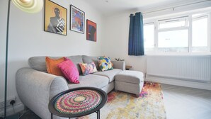 A modern living room with a grey sectional sofa, colorful pillows, a woven coffee table, and artwork on the wall. - The Grove BR1, Spacious 2 Bedroom Flat Greater London (Bromley)