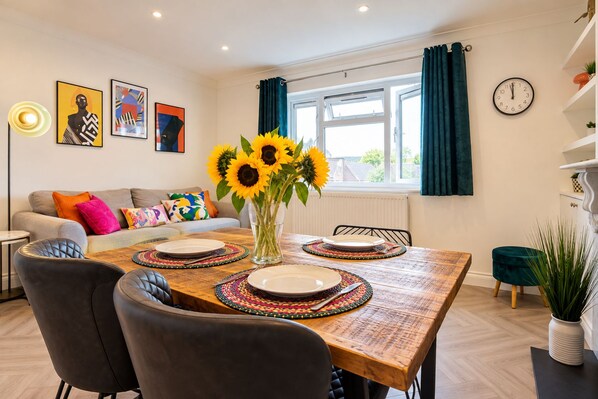 A dining area with a wooden table set for four, surrounded by black chairs. There is a vase with sunflowers on the table, and the room has a modern aesthetic with artwork on the walls and a window with curtains.