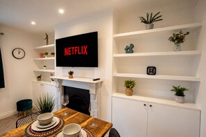 A modern living room with a fireplace, a mounted television displaying 'NETFLIX,' and a dining area with a table set for two. - Stylish 2-Bed Bromley Apartment Short Walk to Town Centre, Spacious 2 Bedroom Flat Greater London (Bromley)