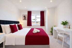 A hotel room with a bed, a desk, a chair, a window with curtains, a plant, and a framed picture on the wall. - Stylish 2-Bed Bromley Apartment Short Walk to Town Centre, Spacious 2 Bedroom Flat Greater London (Bromley)