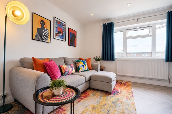 A modern living room with a grey sofa, colorful cushions, a patterned rug, and artwork on the wall.