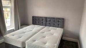 A twin bed with a grey upholstered headboard in a room with light grey walls and a window on the left. - Spacious & Cozy 3 Bedroom Home in Bexleyheath (Bexleyheath)
