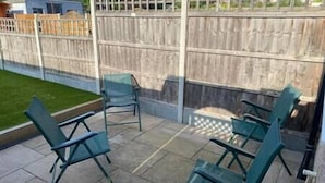 A patio area with four green metal chairs and a small table, enclosed by a wooden fence and a glass partition. - Spacious & Cozy 3 Bedroom Home in Bexleyheath (Bexleyheath)