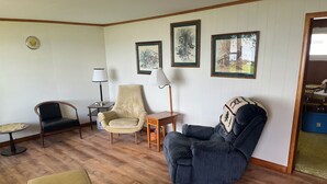A room with wooden flooring, a beige armchair, a dark blue armchair, a small wooden table, and a lamp.