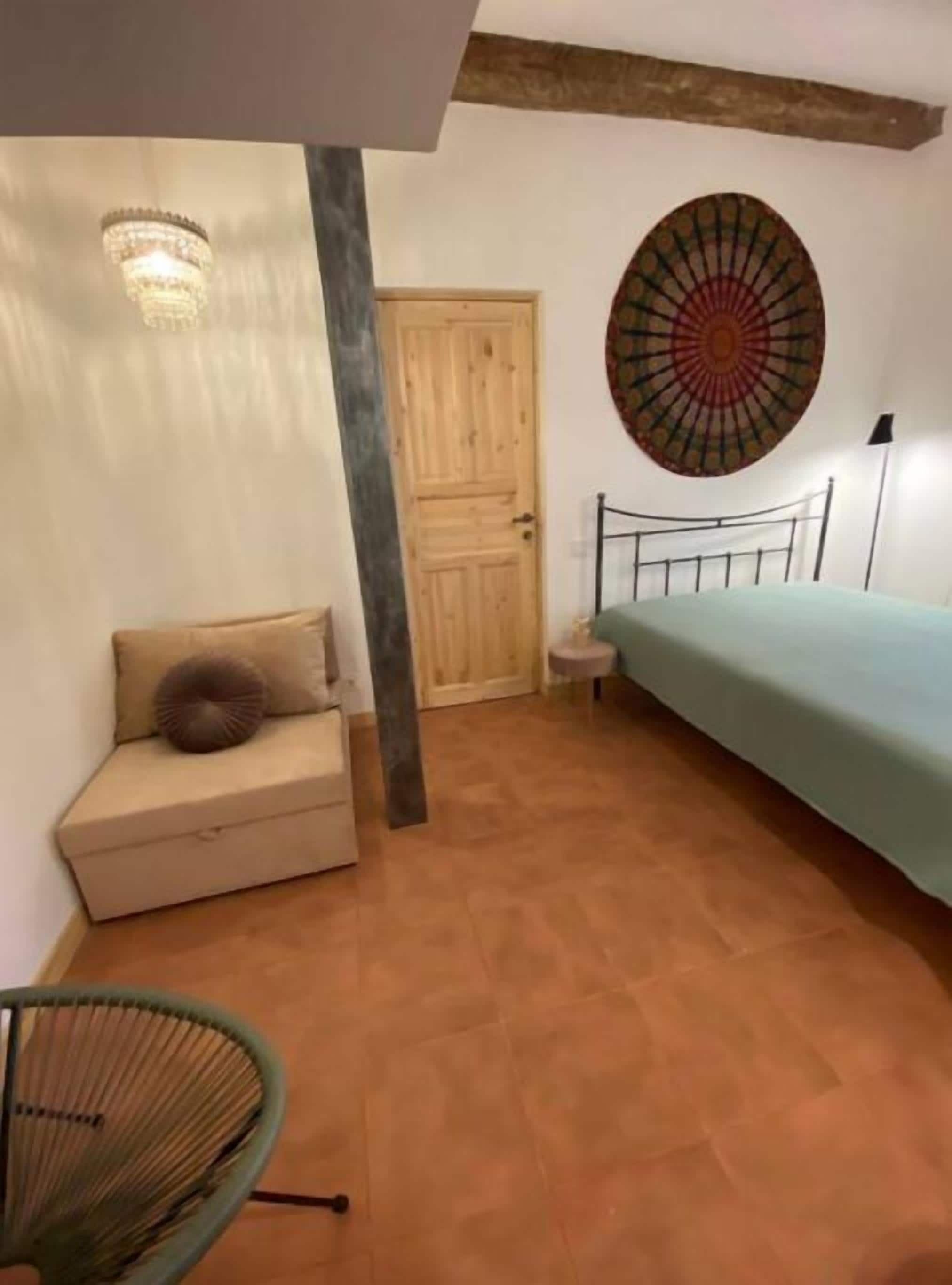 A bedroom with a bed, a wooden door, a wall-mounted artwork, a small table, and a chair.