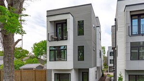 A modern two-story residential building with a balcony on the upper floor, large windows, and a wooden fence in the background.
