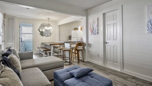 A modern living room with a grey sofa, a blue ottoman, and a dining area with a wooden table and chairs. - Seashore Condo Hotel Two Bedroom Suite, Spacious Fire Island Escape, Near Ferry - With Bay Views and Steps from the Beach. (Ocean Bay Park)