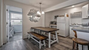 A modern kitchen with a wooden table and bench, a hanging light fixture, and a refrigerator. - Seashore Condo Hotel Two Bedroom Suite, Spacious Fire Island Escape, Near Ferry - With Bay Views and Steps from the Beach. (Ocean Bay Park)