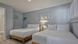 A bedroom with two beds, a radiator, a painting of sailboats, and a window with blinds. - Seashore Condo Hotel Two Bedroom Suite, Spacious Fire Island Escape, Near Ferry - With Bay Views and Steps from the Beach. (Ocean Bay Park)